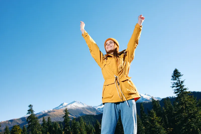 Женщина с рыжими волосами, туристка в жёлтом дождевике и кепке, стоит в горах, подняв руки вверх от счастья, любуясь видом на заснеженные горы