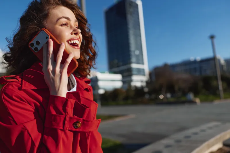 A joyful woman in a red coat chats on a smartphone outdoors in an urban plaza, sunlight bright, modern buildings in the background, light breeze, casual chic mood.