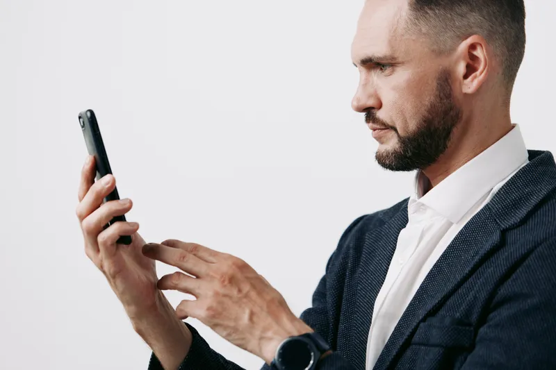 A focused business professional man stands against an isolated colored background, examining a smartphone with calm determination, confident posture, and thoughtful expression.