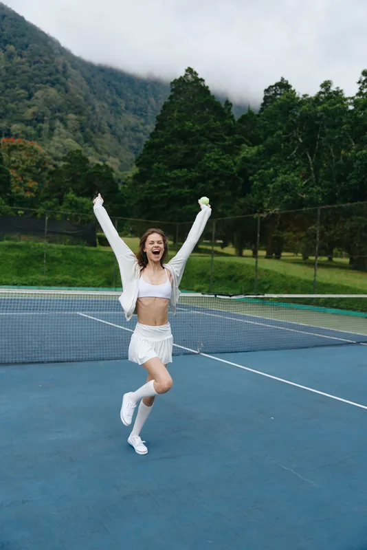 Happy female tennis player celebrating victory on the court with a joyful expression, wearing a stylish white athletic outfit amidst a lush green background