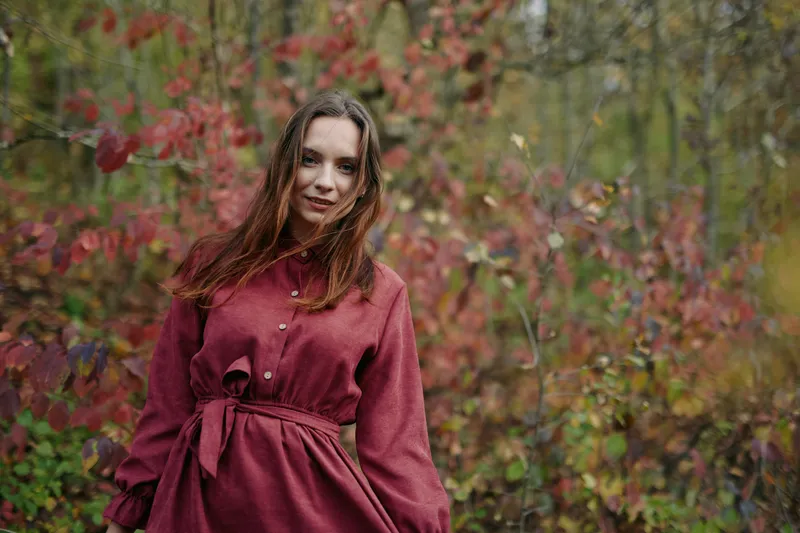 A young woman in a burgundy dress stands among autumn foliage, smiling naturally with braided hair and soft natural light, conveying an authentic, genuine portrait and candid presence in nature.