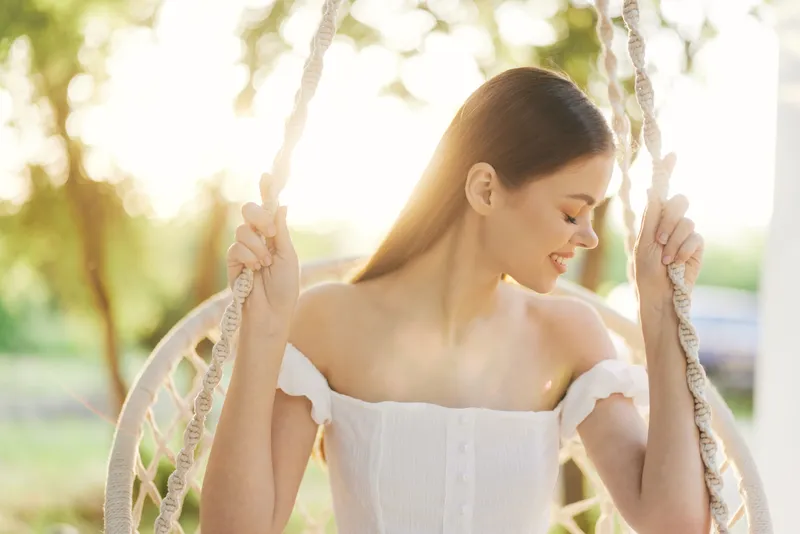 Serene young woman in off shoulder dress swinging gently, enjoying nature s beauty in a sunny garden, embodying tranquility and joy in a vibrant summer setting