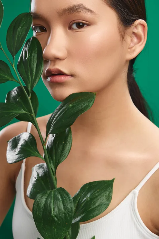 asian woman with smooth skin looks directly at the camera among green leaves, studio portrait with bright light, clean aesthetic, calm mood, contemporary fashion vibe, natural softness, poised