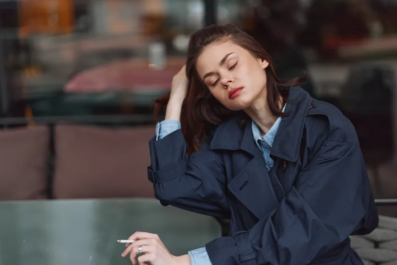 A young woman in a dark coat sits indoors with eyes closed, holding a cigarette, appearing tired and contemplative in a dimly lit cafe setting.