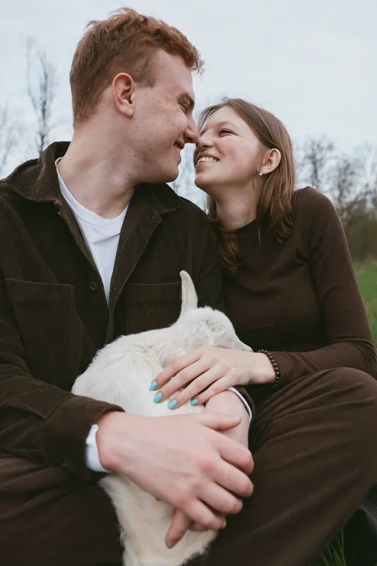 Cheerful couple outdoors, smiling while holding a goat kid, intimate moment, soft natural light, relaxed posture, rustic setting, shared affection, candid lifestyle scene, earthy colors, gentle