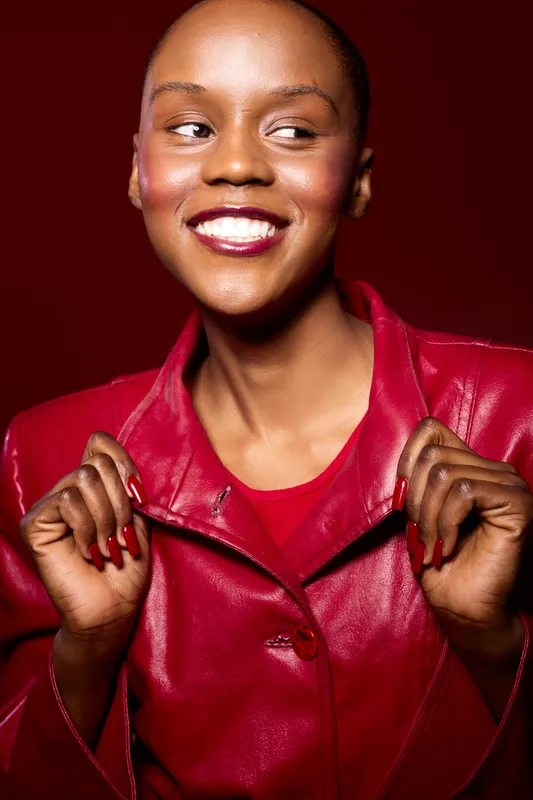 Smiling African woman with dark skin wearing red leather jacket and matching nails looks aside on red background. Concept of fashion, beauty, confidence, and stylish portrait.