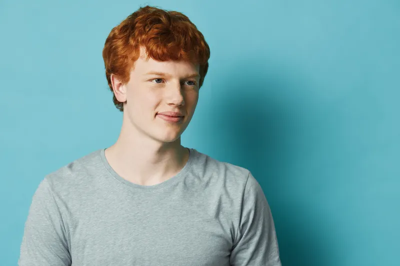 Young man with red hair wearing gray t shirt looking sideways against blue background in studio with natural expression and casual posture.