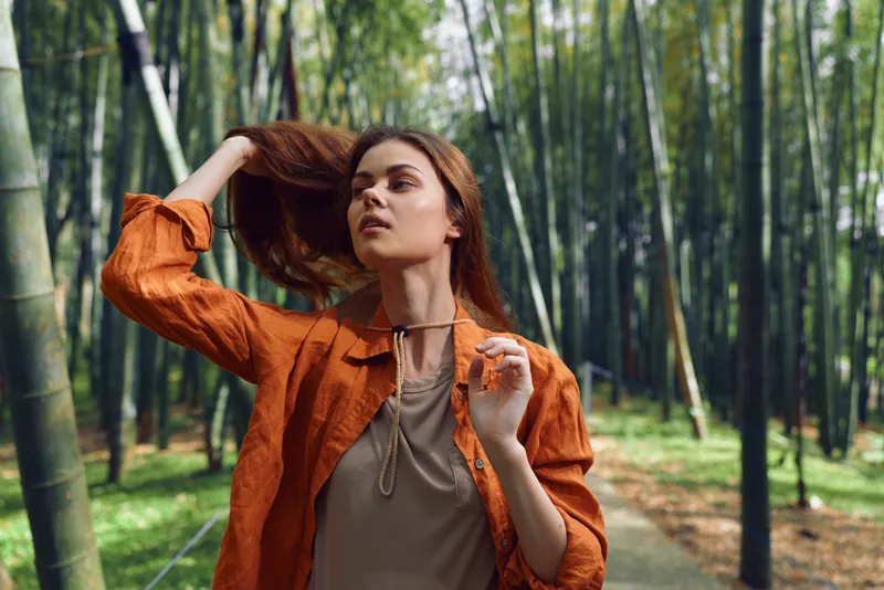 Woman portrait in bamboo forest, fashion model wearing orange jacket and bohemian outfit, walking on a path with hair flipping, serene nature background and soft sunlight.
