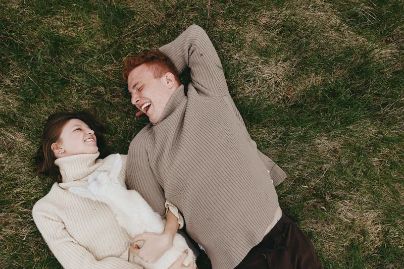 Young couple lying on grass holding a baby goat and smiling at each other in cozy sweaters during a relaxed outdoor moment in nature.