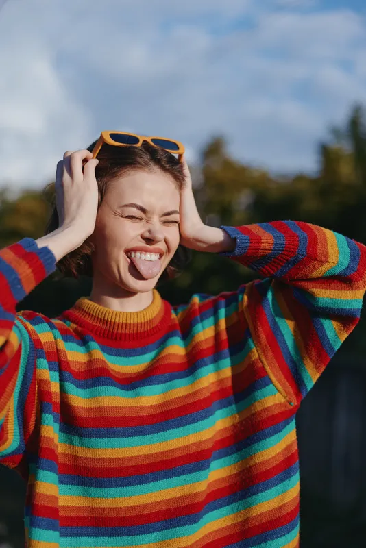 Young woman in her early twenties wearing a rainbow sweater and sunglasses on head, playful expression sticking tongue out, casual fashion portrait outdoors in sunny park.