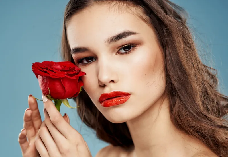 Женщина держит в руке красный цветок на синем фоне, на её лице макияж