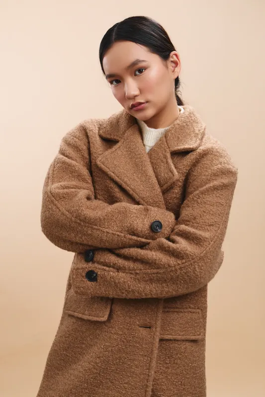 Asian woman stands in a neutral studio wearing a brown wool coat, arms crossed, composed expression, exuding confidence, sophistication, and a modern fashion poised mood.