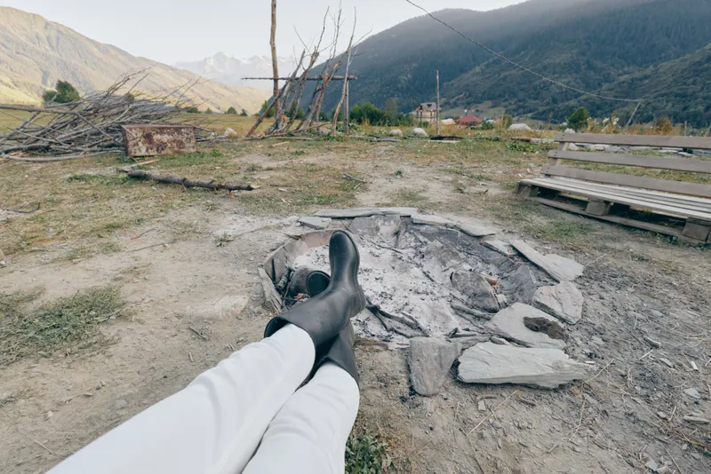 Boots, legs, campfire, ashes, mountain, bench, countryside, relaxation — person in rubber boots relaxing with legs outstretched toward an empty fire pit in a rustic mountain valley outdoors.