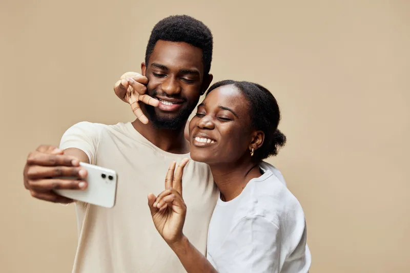 Happy young African couple taking a selfie, casual clothing, joyful expressions, neutral background, perfect for social media and relationship concepts