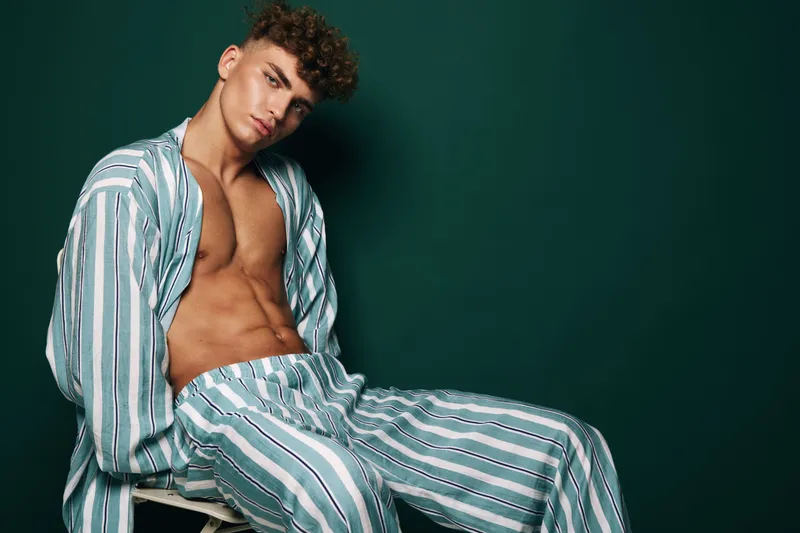Young man with curly hair wearing striped pajamas sitting on stool against dark green background in studio for fashion and lifestyle portrait.