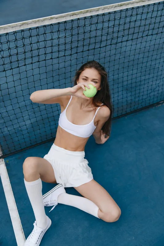 Young woman enjoying a green apple while sitting on a tennis court, wearing a white sports bra and shorts, exuding health and vitality