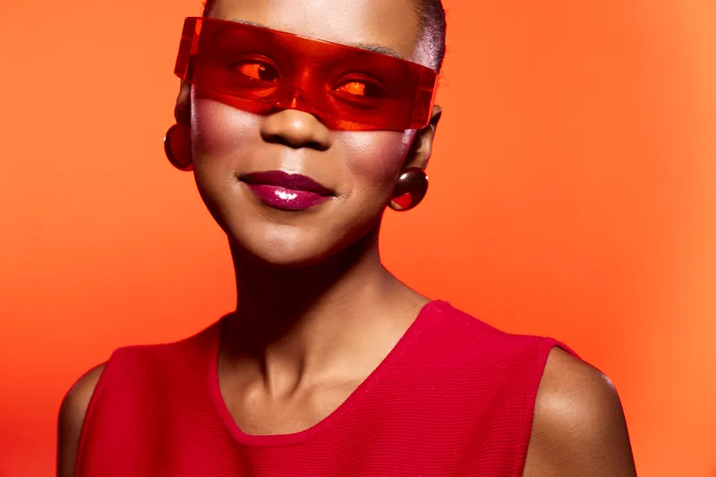 Portrait of a confident black woman with dark skin wearing red sleeveless top and stylish red sunglasses against vibrant orange background. Concept of fashion, beauty, and empowerment.