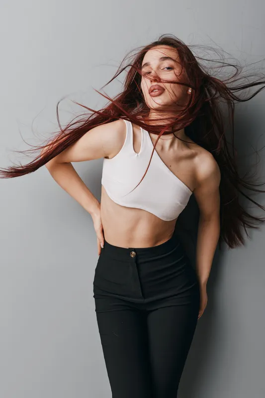 Young woman with long hair wearing white crop top and black pants posing with hand on hip against gray background. Fashion model with flowing hair and confident expression in studio.