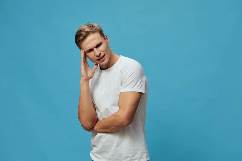 Boy experiencing headache, holding head, suffering from pain, young man with discomfort, isolated on solid blue background, casual clothing, health problem concept, emotional stress, studio portrait, 
