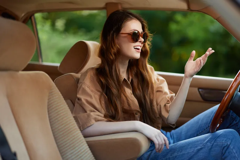 Young woman with long hair enjoying a drive in a vintage car on a sunny day, wearing sunglasses and a relaxed outfit.