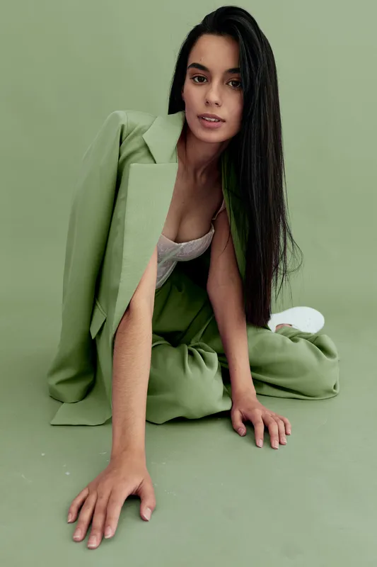 young woman with long dark hair wearing green suit posing on the floor against a light green background, stylish contemporary fashion look