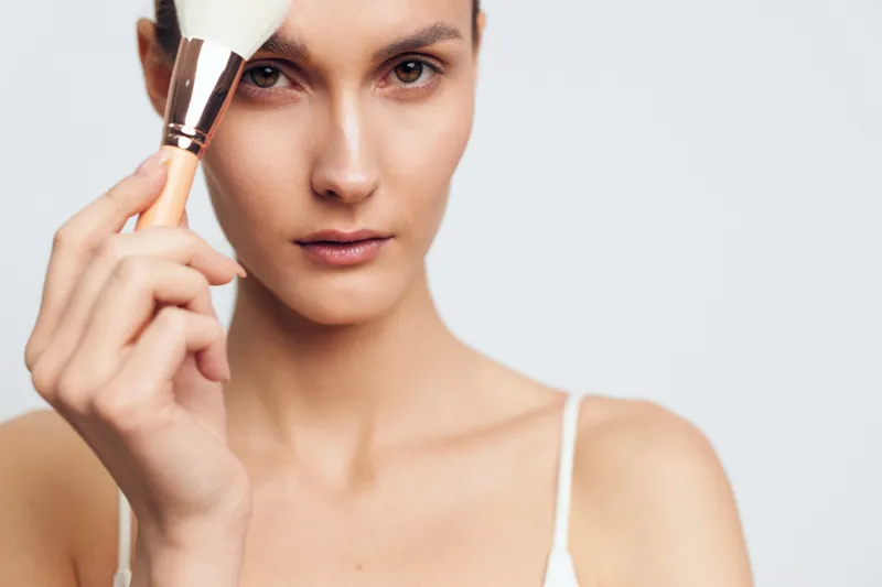 Young woman applies makeup with brush on face closeup portrait natural skin tone white background beauty concept skincare routine