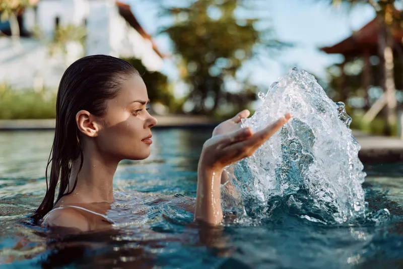 Young woman enjoying outdoor pool, holding water splash in hand, wet hair, calm expression, tropical background, sunny day, natural lifestyle moment