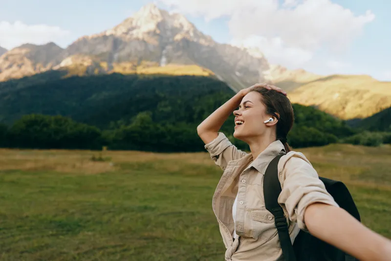 Woman outdoors, arms spread, smiling in a meadow with distant mountains. Casual travel lifestyle, nature, and freedom vibe, capturing joyful exploration and wellness.