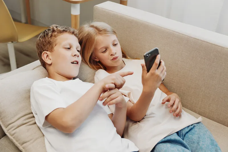 Two children lying on a sofa using smartphone together, engaged in digital activity. Casual home environment with natural light.