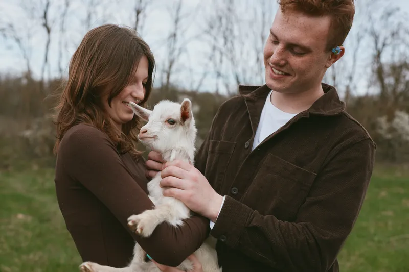 A couple outdoors holds a goat kid with smiles, capturing a wholesome lifestyle moment in a sunny field, showing affection, companionship, nature, and farm life with a playful animal.