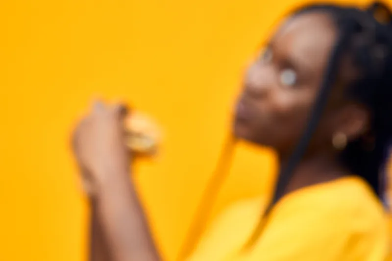 Young woman enjoying a burger in vibrant yellow attire against a bright yellow background, expressing joy and satisfaction in a modern, trendy setting