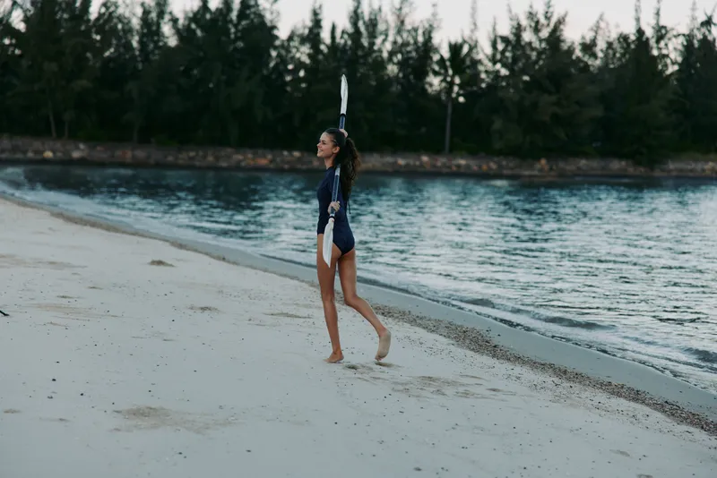 A woman walks along a sandy beach with a surfboard raised above her head, ready for a ride as calm water and a pine forest backdrop frame the scene.