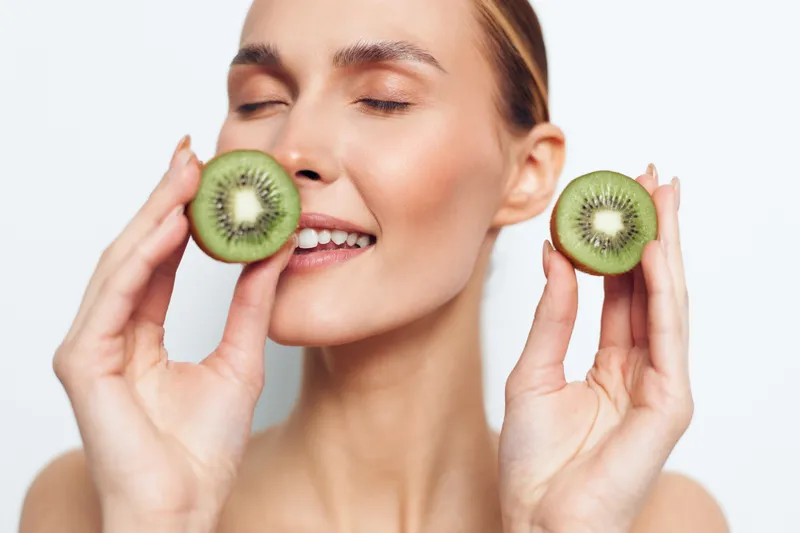 Young woman with clear skin holding two halves of fresh kiwi fruit near her face smiling with closed eyes on white background. Concept of healthy lifestyle and natural beauty.