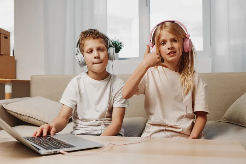 Two children wearing headphones enjoy music together at home, sitting on a couch with a laptop in front of them. The girl shows a thumbs up while the boy listens with eyes closed.
