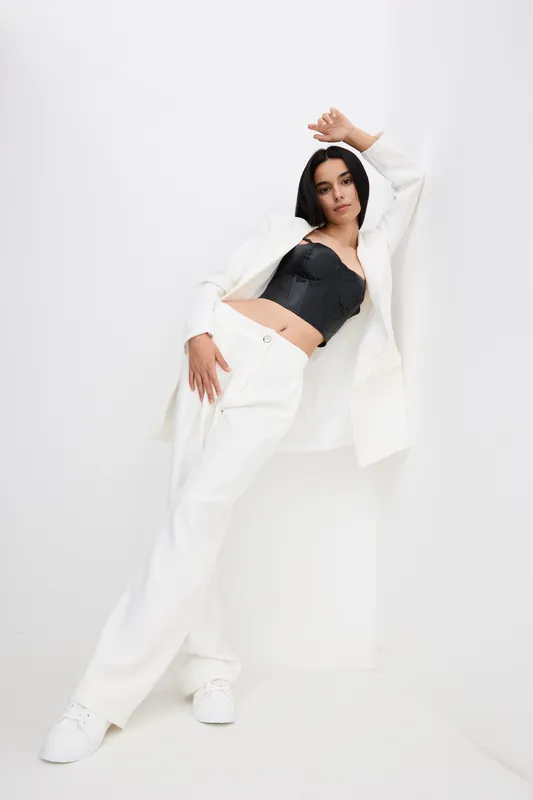 Stylish woman in white wide-leg pants and jacket posing confidently against a white corner wall, wearing a black corset top and white sneakers with a modern minimalist vibe.