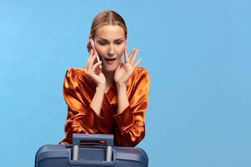 Surprised woman in orange blouse talks on phone while holding blue suitcase against blue background. Concept of travel, communication, and unexpected news or plans.