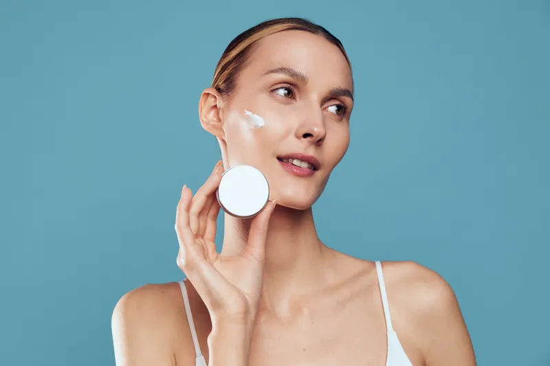 Woman applies face cream with cotton pad, skincare beauty routine for hydration and smooth skin, calm expression on blue background, self care concept for cosmetic care.
