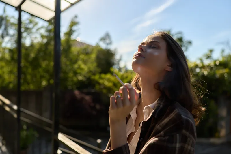 A woman outdoor in a plaid jacket looks upward, cigarette in hand, bathed in warm sunlight. A contemplative lifestyle portrait capturing thoughtful, casual style and relaxed moments in natural light