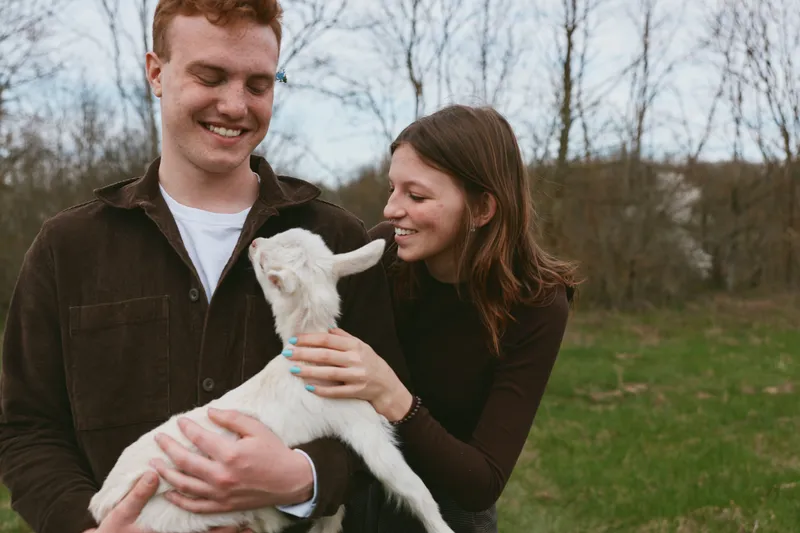 Happy couple outdoors with a playful goat kid, smiling and gentle, cuddling the animal in a sunny field, capturing a warm lifestyle moment of friendship