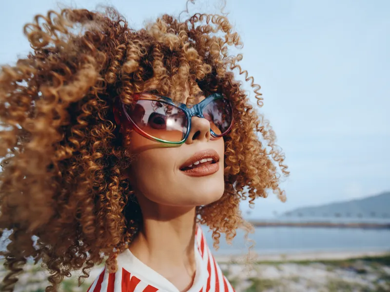 Portrait of a young woman with curly hair and stylish sunglasses posing outdoors by a sunny coastline, capturing a bold, confident, and playful mood.