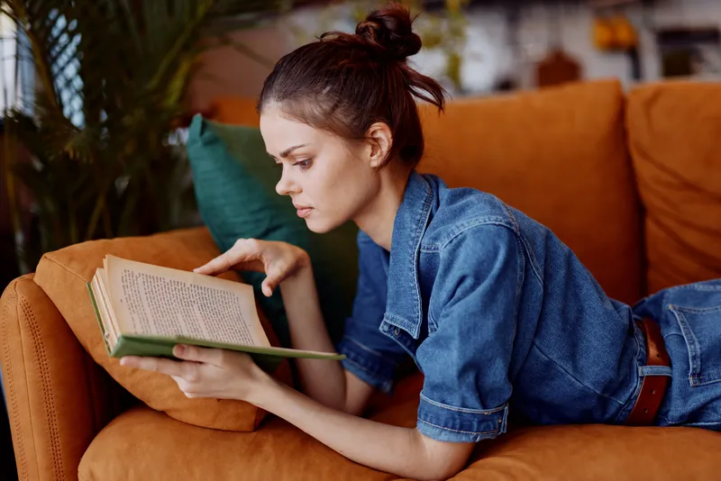 Женщина в джинсовой рубашке читает книгу, сидя на оранжевом диване в уютной гостиной
