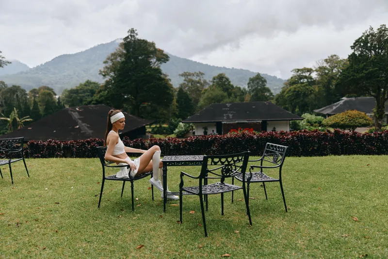 A serene young model in activewear poses thoughtfully outdoors amidst a lush garden with vibrant greenery and distant mountains, capturing a moment of reflection and tranquility