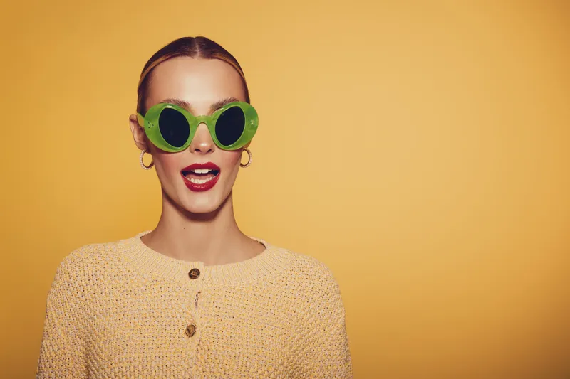 Young woman with green round sunglasses and beige knitted sweater poses against yellow background. Fashion portrait with trendy accessories and casual style. Concept of summer and fun.