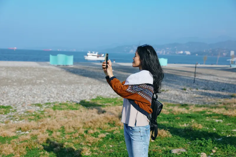 Selfie of a woman on a sunny beach, capturing the sea, distant boats, casual outfit, wind in hair, grass and shoreline in frame, outdoor lifestyle and travel moment photography