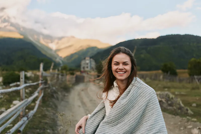 Bright portrait of a smiling woman outdoors wrapped in a shawl, standing along a rural road with mountains and blue sky in the background, capturing carefree nature