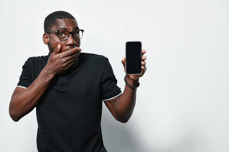 African American man with dark skin and glasses showing smartphone with blank screen and covering mouth in surprise or shock on white background.