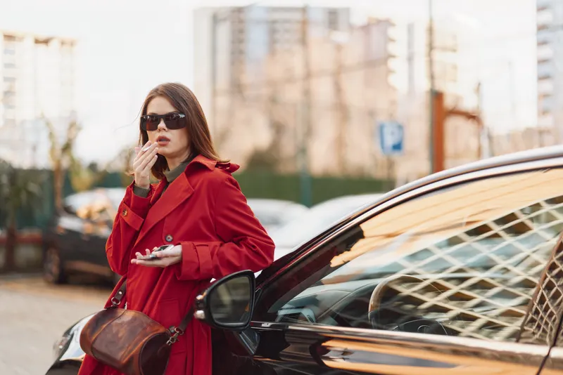 Cheerful woman in a red coat beside a parked car, sunglasses on, talking on the phone, urban street scene, warm sunlight, contemporary fashion, candid moment.