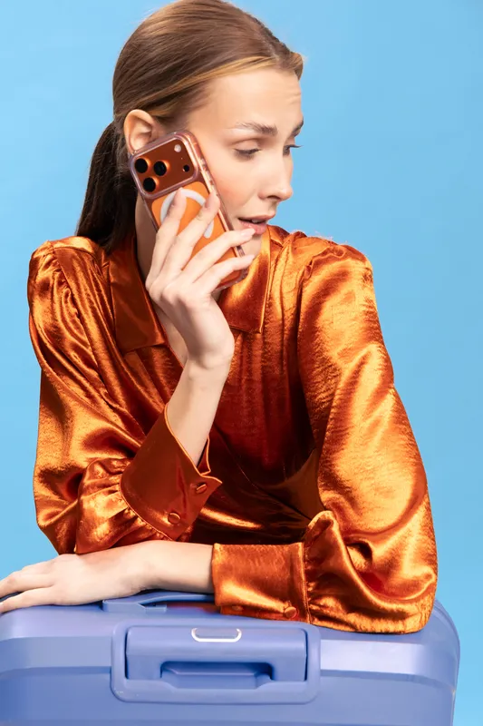 Young woman in orange satin blouse talks on smartphone while leaning on blue suitcase. Studio portrait with blue background. Concept of travel, communication, and fashion.