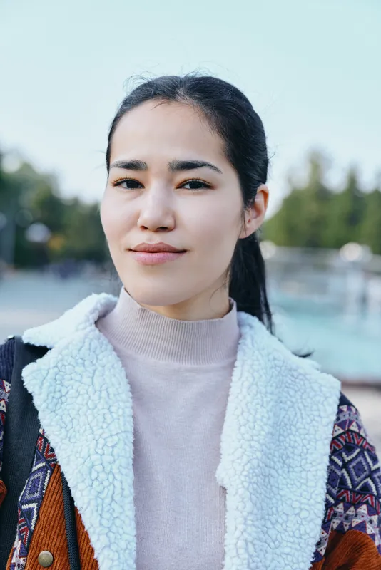 Woman wearing a cozy shearling jacket in an outdoor setting, calm expression, soft daylight, casual style, street fashion atmosphere, lens flare and blurred background.