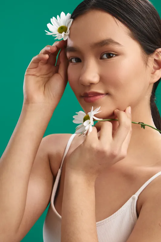 asian woman with a gentle smile poses with flowers against a vivid green studio backdrop, showcasing a fresh and confident aesthetic that conveys warmth, charm, and modern poise in a bright,